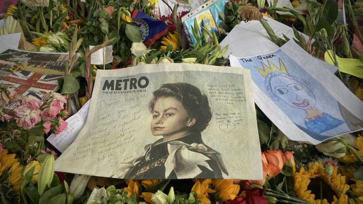 Colorful, decaying flower bouquets. In the middle, a copy of the London Metro with Queen Elizabeth II on the front page. 